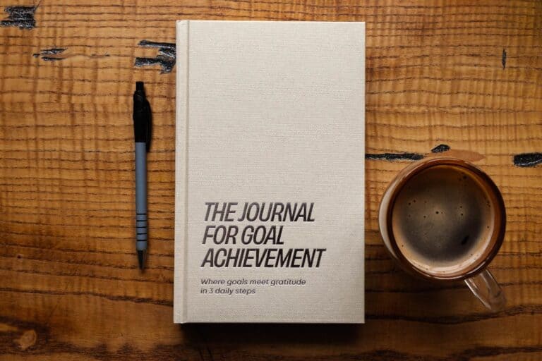 "A hardcover journal titled 'The Journal for Goal Achievement' placed on a wooden desk with a pen and a cup of coffee, symbolising focus, consistency, and daily goal-setting. Learn how to stay consistent with your goals using structured journaling.