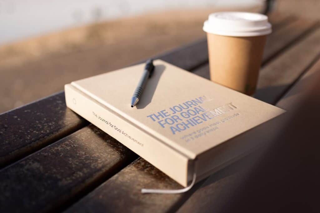 The Journal for Goal Achievement on a park bench with a pen and takeaway coffee cup — a daily tool to help you stick to your goals.