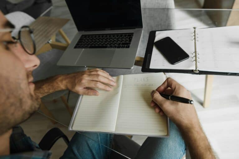 Person writing plans in a paper journal beside a laptop and planner during hybrid work