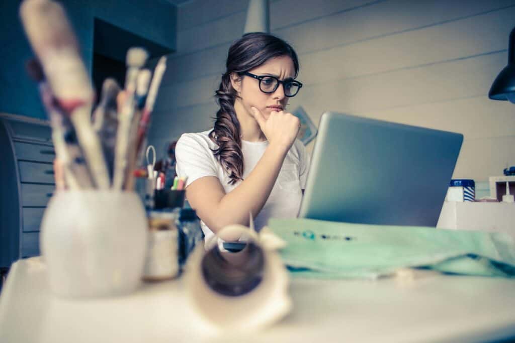 Thoughtful woman staring at laptop, surrounded by art supplies