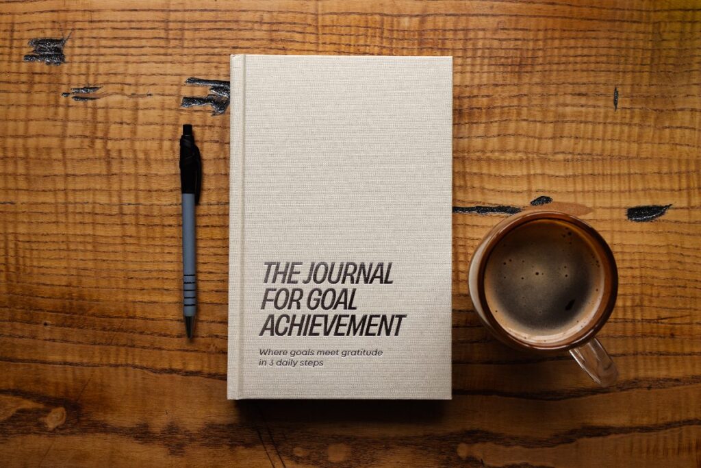The Journal for Goal Achievement on a wooden desk with a pen and coffee for goal-setting and weekly reflection.